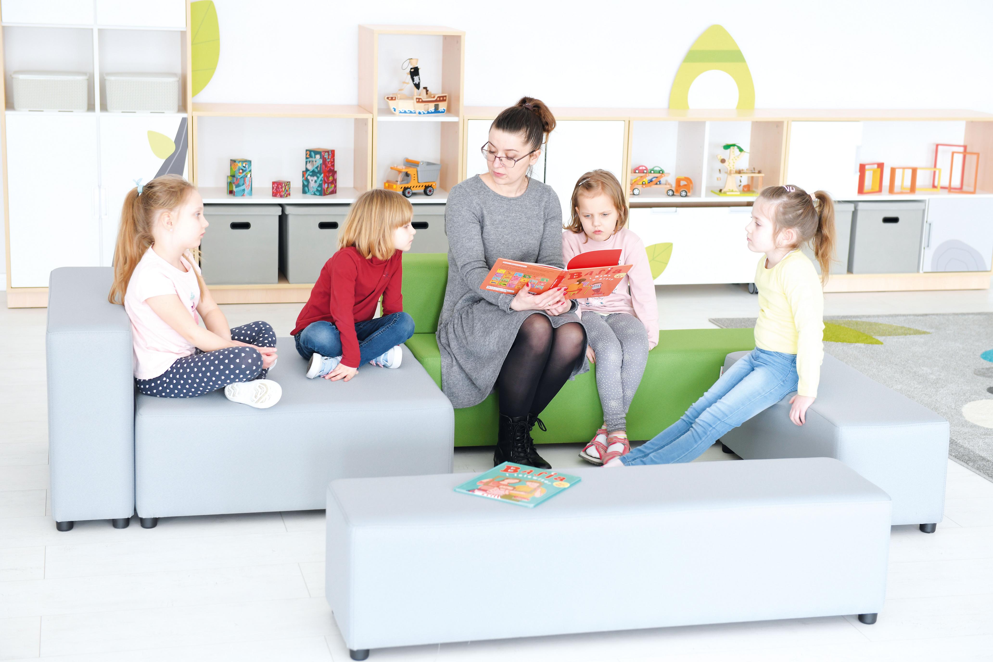 Kinder und Erzieherin auf modularen Sitzlandschaften, lesen Buch.